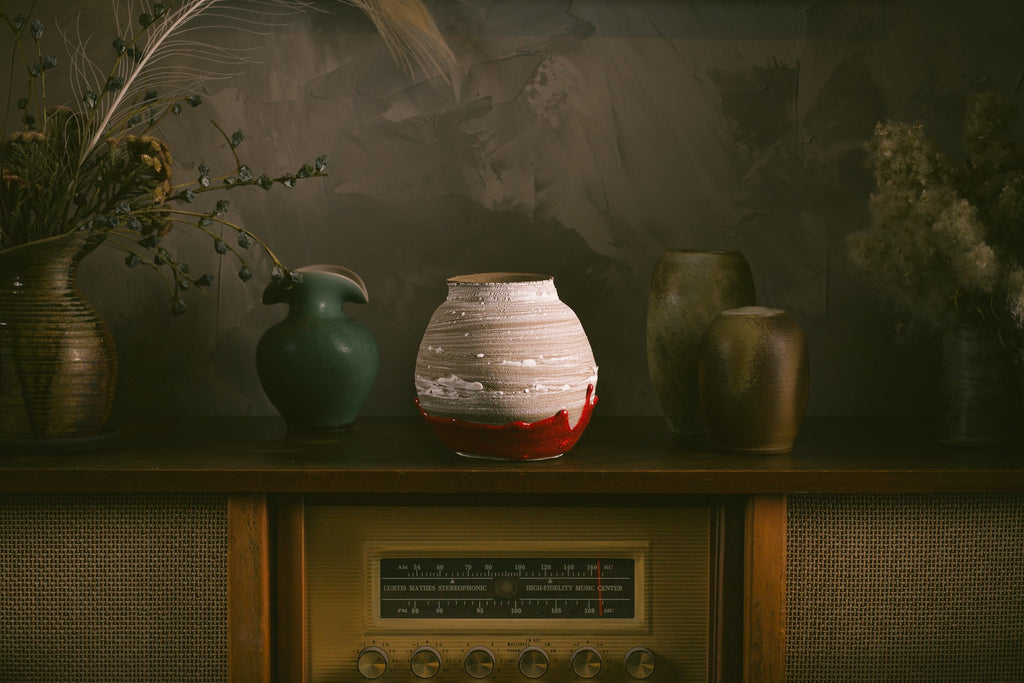 Painting of a white and red vase in the middle, with a turquoise vase to the left and two brown vases on the right. The vases are all on top of a counter with a radio in the middle. There are vases of flowers on either end of the photo.